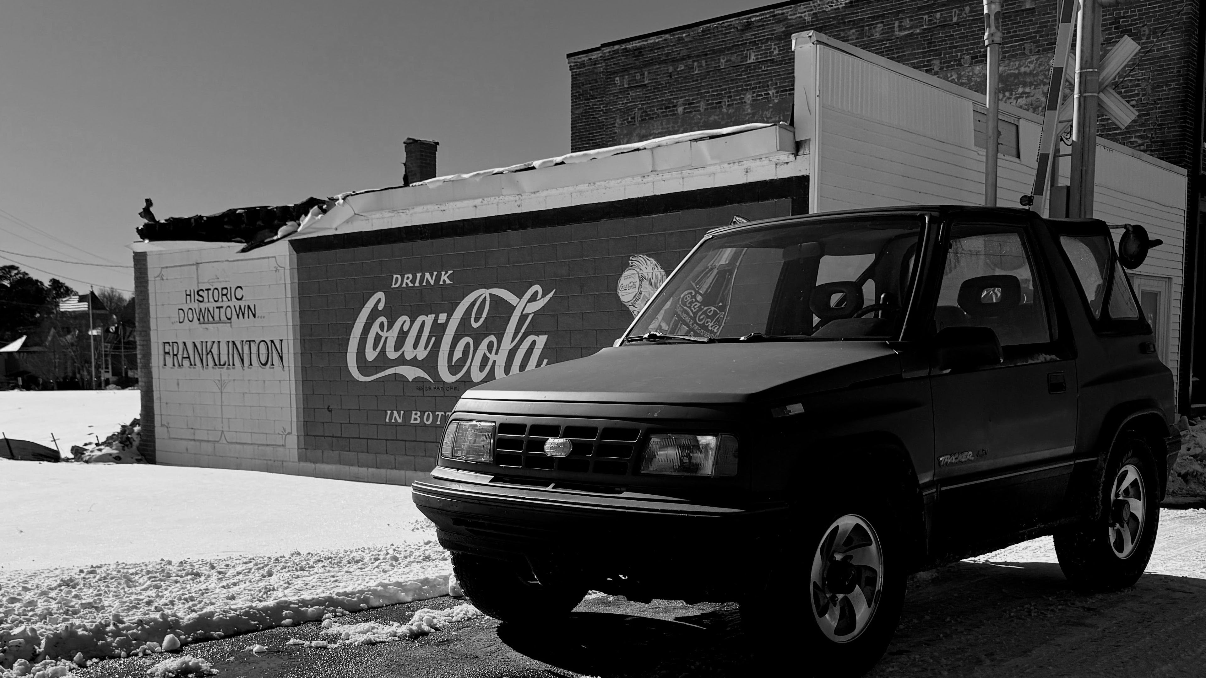 Vintage SUV parked in front of a Coca-Cola truck with a snowy ground and building in the background.